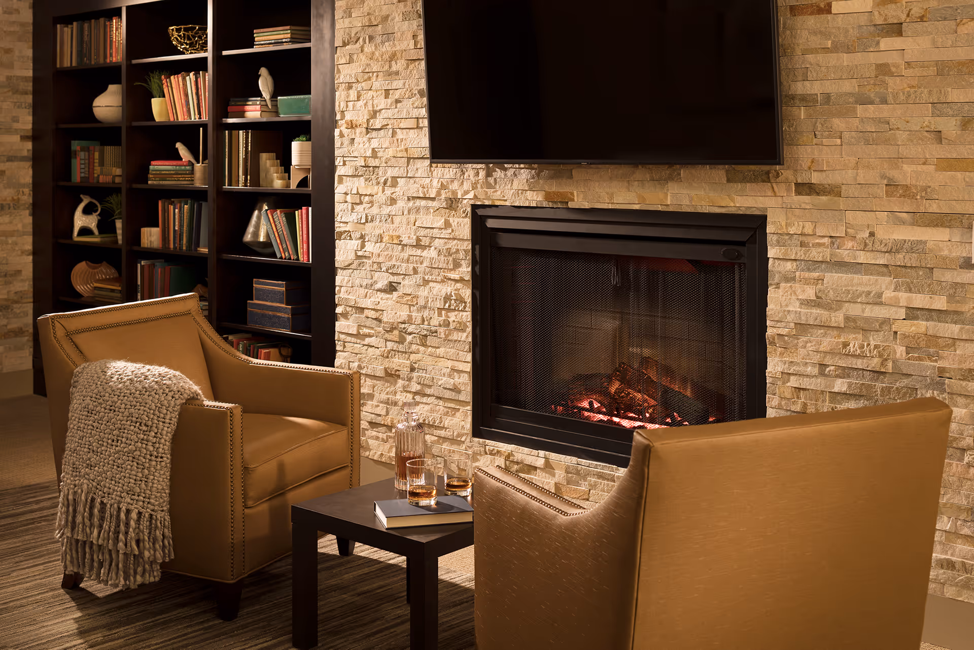 Cozy living room area with two tan armchairs facing a modern stone fireplace with a flat-screen TV mounted above it. A small dark wooden table between the chairs holds a decanter and two glasses, along with two books. A built-in dark wood bookshelf filled with books and decorative items is visible in the background.