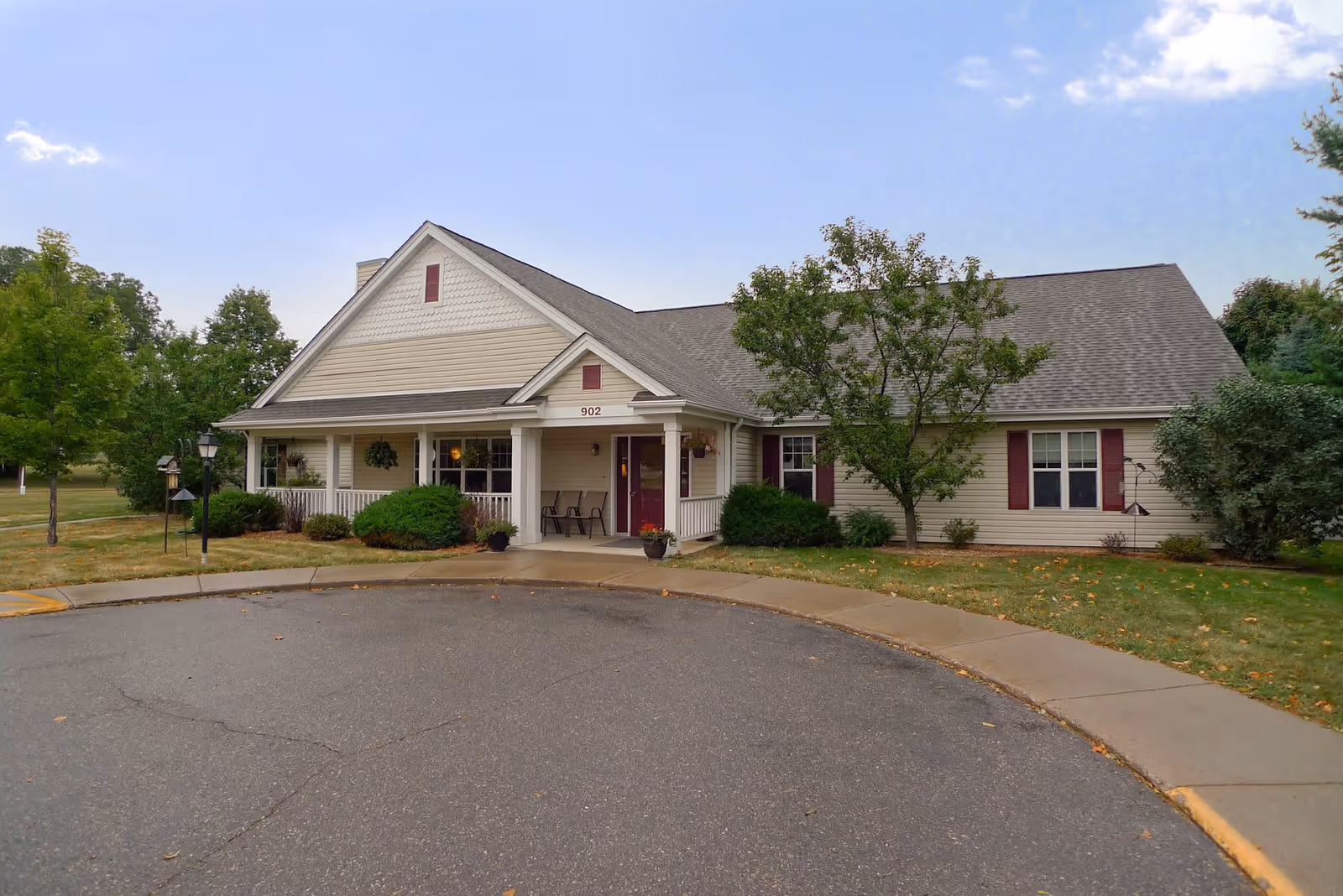 Exterior view of a single-story senior living facility building with beige siding, a gray shingled roof, and a covered front porch with chairs. The building is surrounded by green bushes, trees, and a lawn, with a paved circular driveway in front under a partly cloudy sky.