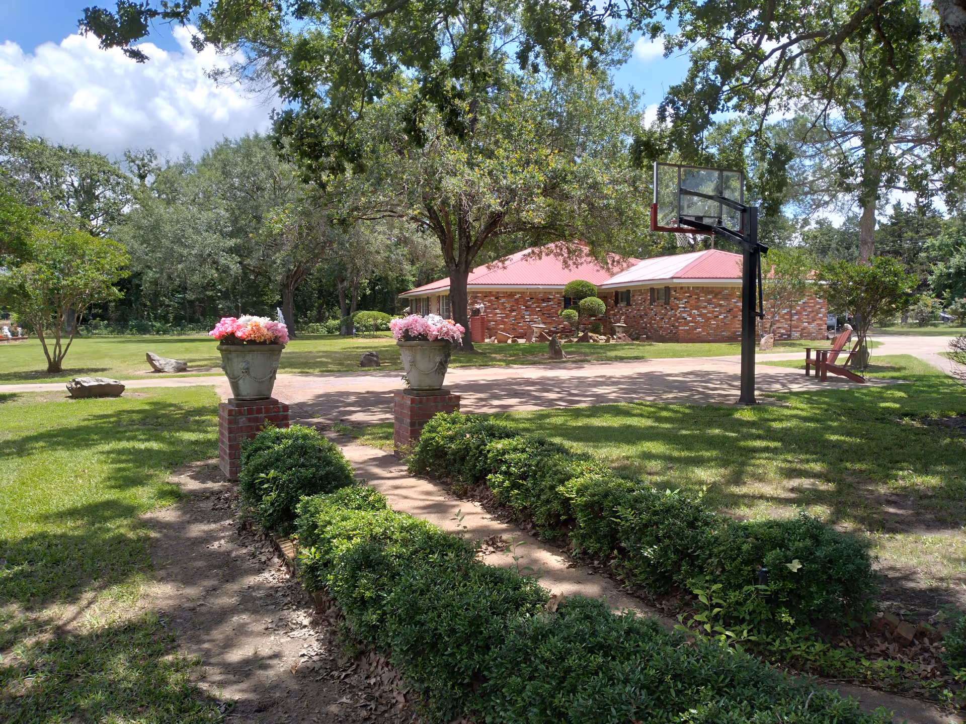 Outdoor view of a senior living facility with a brick building featuring a red roof in the background. The foreground shows a garden area with trimmed bushes, two flower pots with pink flowers on brick pedestals, a basketball hoop, and a wooden chair under the shade of large trees.