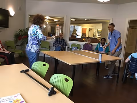 A group of elderly people and a caregiver playing table tennis in a common room with green chairs and wooden floors. The room has large windows into an adjacent area and a TV mounted on the wall.