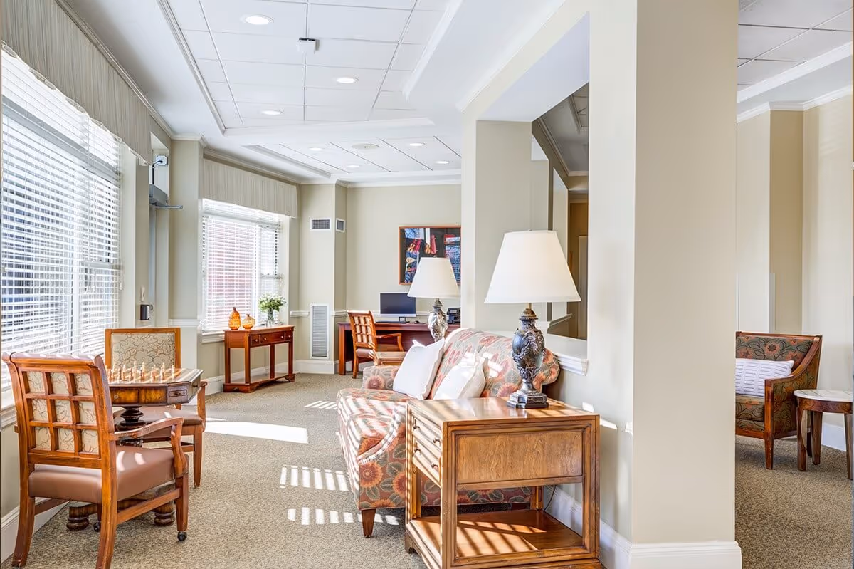 Sunlit communal sitting area with a patterned sofa, wooden side tables and lamps, armchairs, and a chessboard by large windows.