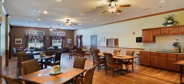 A spacious dining room with multiple wooden tables and chairs arranged neatly. The room features hardwood flooring, ceiling fans with lights, large windows with curtains allowing natural light, and a kitchenette area with cabinets, a countertop, and a coffee machine. Small flower arrangements are placed on the tables.
