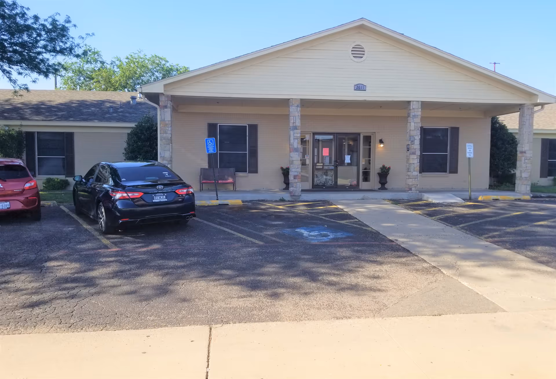 Front exterior view of Georgia Manor Nursing Home building with a covered entrance supported by stone pillars, two parked cars in front, and a clear blue sky.