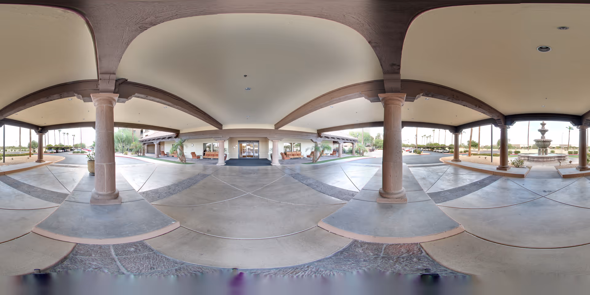 Covered entrance area of The Forum at Desert Harbor with large stone columns supporting the roof, a circular driveway, and a fountain visible to the right. The entrance doors to the building are straight ahead, with benches and landscaping around the area.