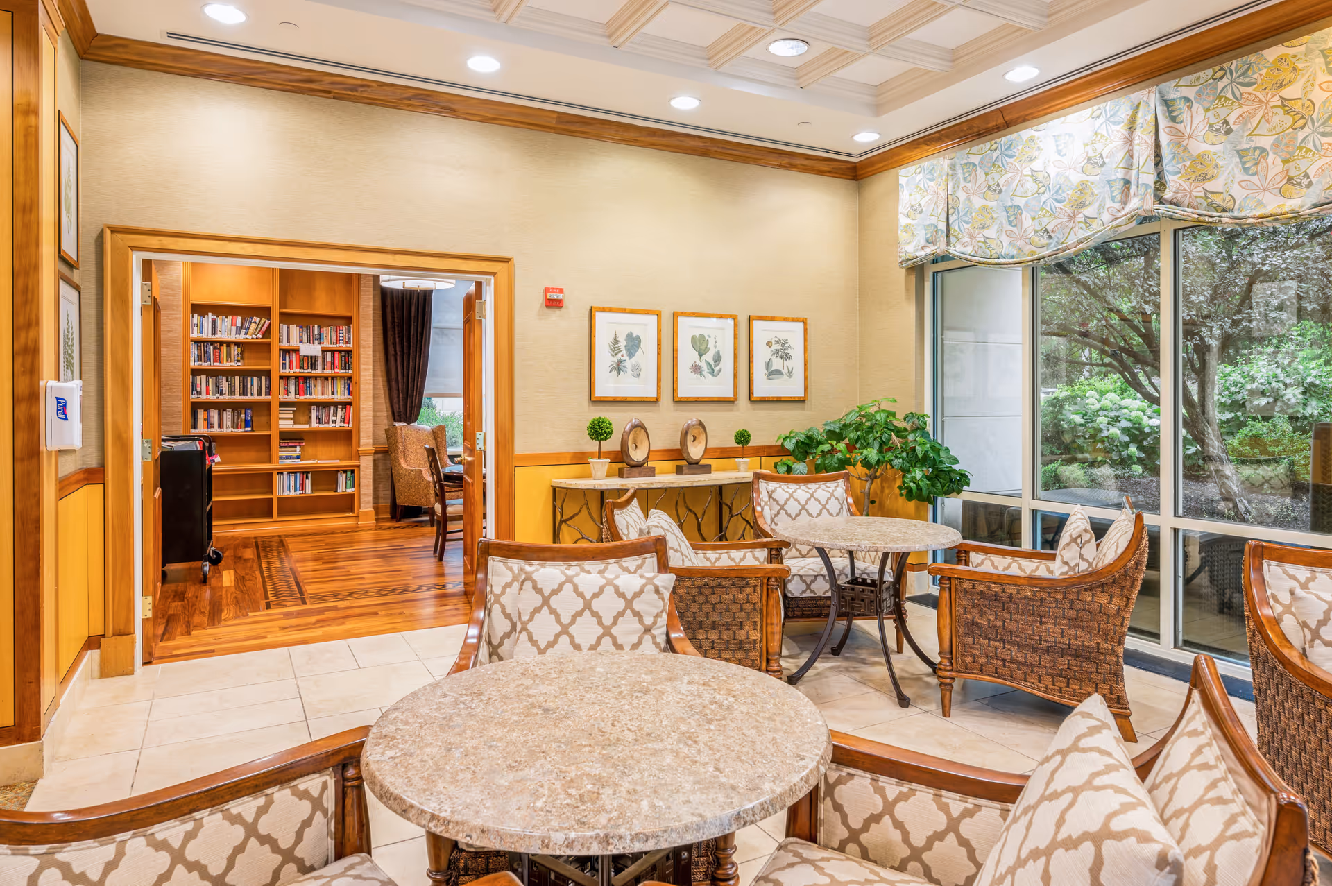A cozy seating area with round tables and cushioned wicker chairs in a senior living facility. The room has large windows with floral curtains overlooking a garden. In the background, there is an open doorway leading to a library with wooden bookshelves filled with books and a wooden floor.