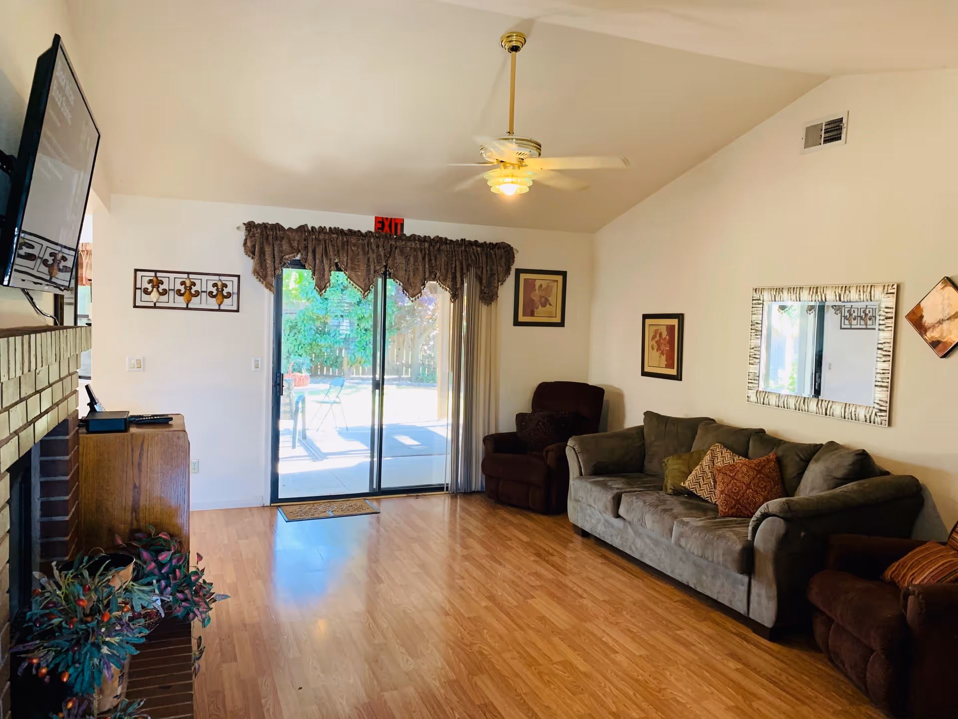 A cozy living room with wooden flooring, a gray sofa with multiple cushions, a brown armchair, a large mirror on the wall, framed artwork, a ceiling fan with light, a sliding glass door with curtains leading to an outdoor patio, and a fireplace with a TV mounted above it.