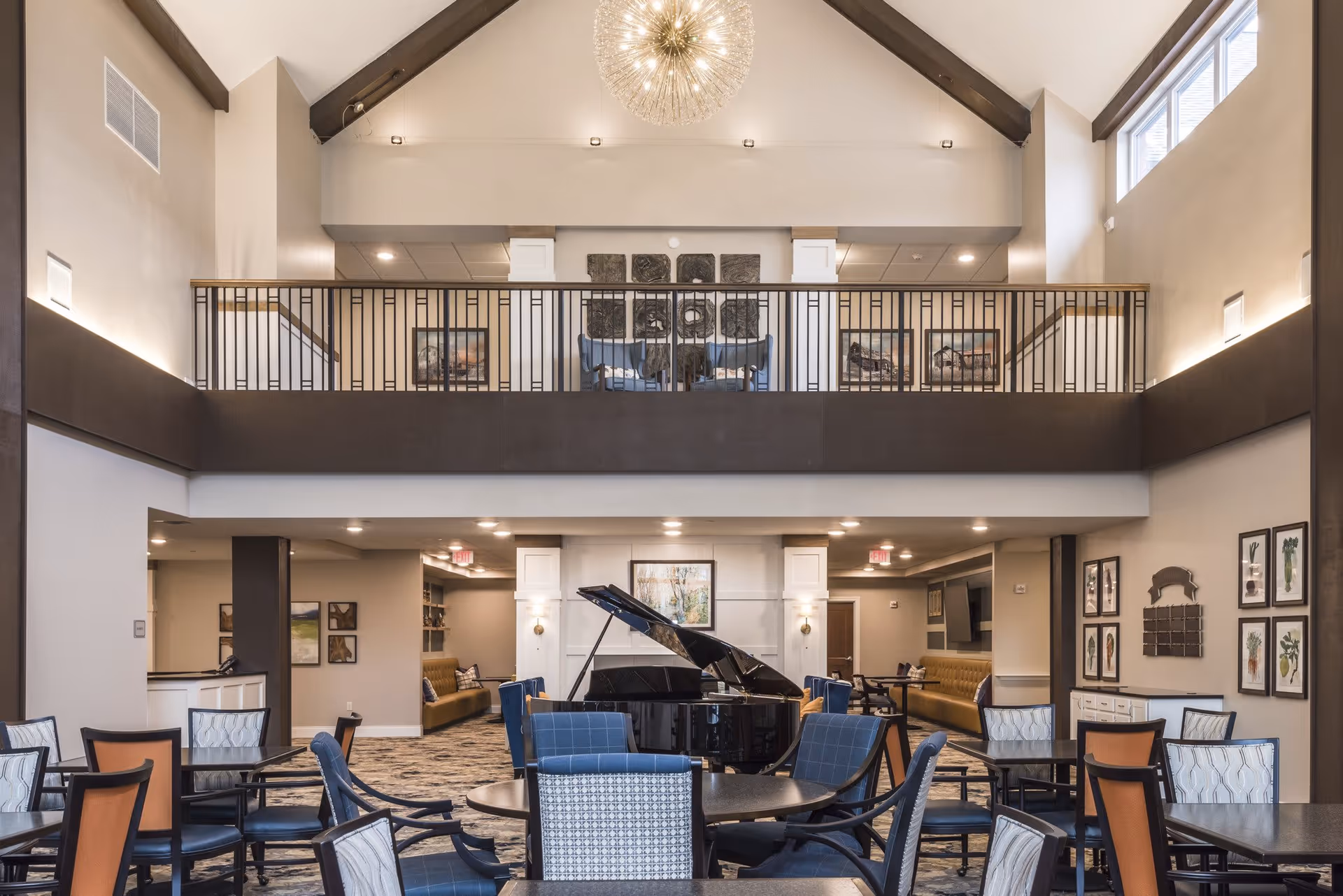 A spacious and well-lit common area in a senior living facility featuring a grand piano in the center, surrounded by multiple tables and chairs with patterned upholstery. The room has a high ceiling with a modern chandelier, a balcony with railings overlooking the space, and framed artwork on the walls.