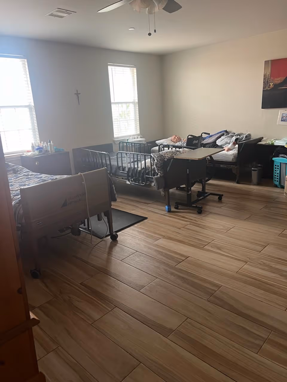 Shared assisted living bedroom with multiple hospital-style beds, a resident lying in one bed, windows, and wood-look tile flooring.