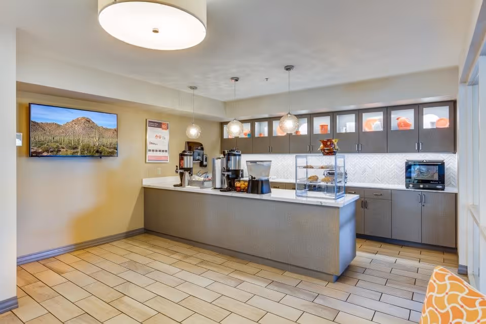 A modern kitchen area in a senior living facility with a long island counter featuring coffee dispensers, a water dispenser, and a display case with snacks. The back wall has cabinets with glass doors on the upper section, displaying orange and white dishes. A microwave is placed on the right side countertop. A flat-screen TV is mounted on the left wall showing a desert landscape. The floor is tiled and there is a round ceiling light fixture above.