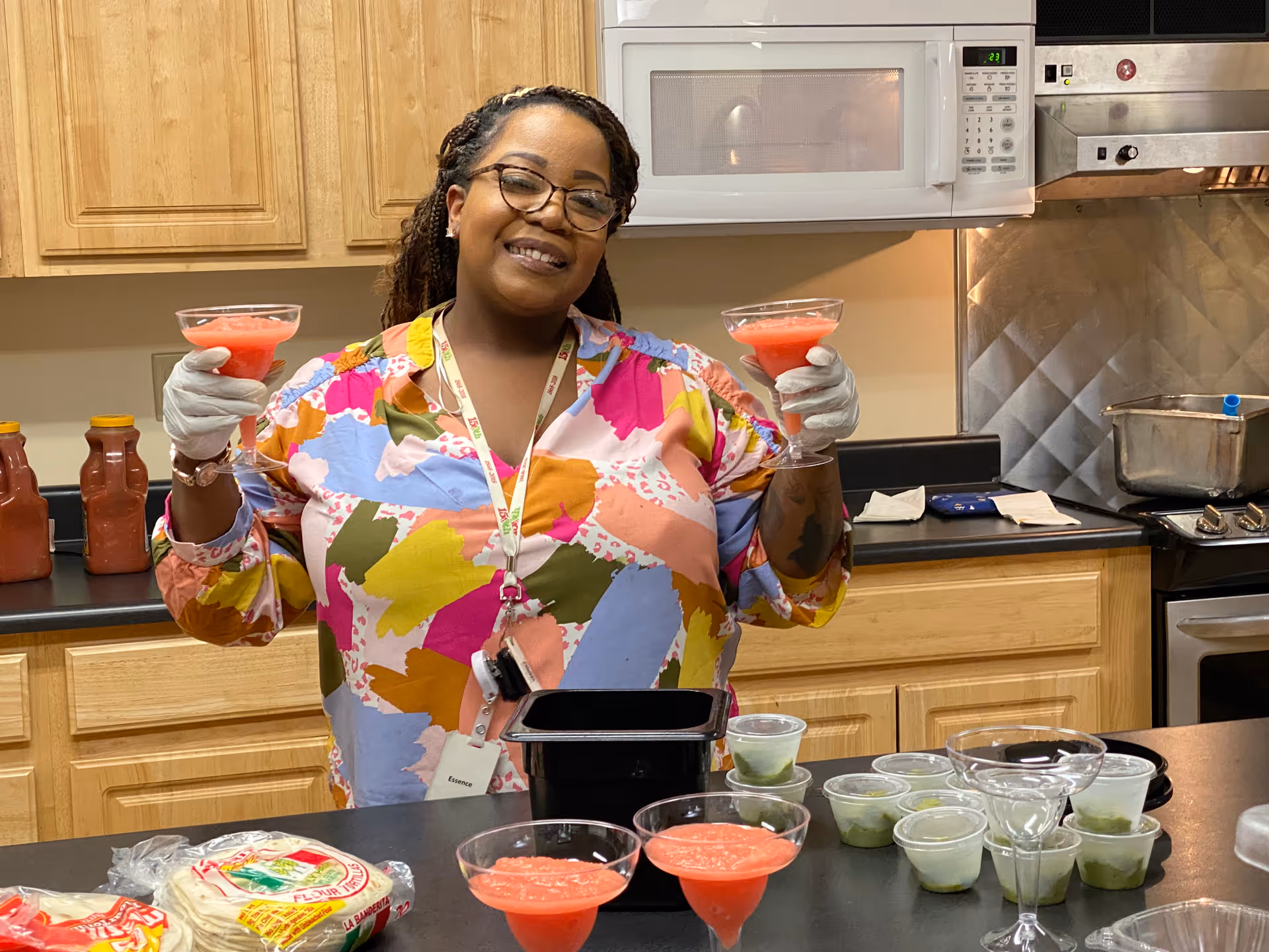 A smiling woman wearing glasses and a colorful blouse stands in a kitchen holding two glasses filled with a red beverage. She is wearing gloves and standing behind a counter with several cups containing green and red liquids, tortillas, and kitchen utensils. Wooden cabinets, a microwave, and a stove are visible in the background.