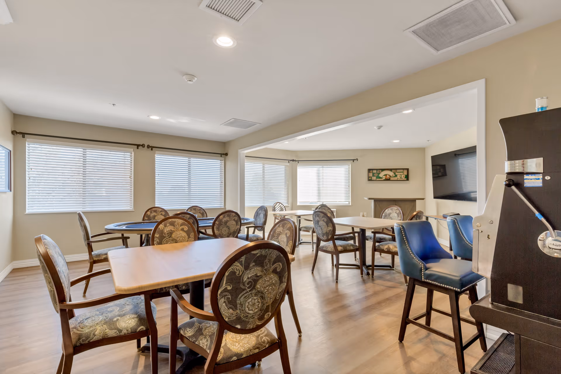 A bright and spacious common area with multiple tables and chairs arranged for seating. The room has large windows with blinds allowing natural light to fill the space. There is a flat-screen TV mounted on the wall and a coffee or beverage machine with blue cushioned chairs nearby. The flooring is wood, and the walls are painted a light beige color.