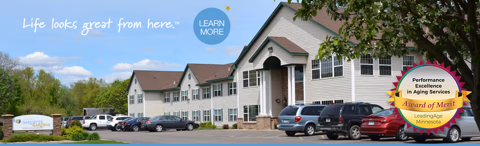 Exterior view of Vista Prairie at Garnette Gardens senior living facility showing a two-story building with multiple windows, a parking lot with several cars, and a sign for Garnette Gardens. The sky is blue with some clouds, and there are trees in the background. There is also a badge indicating an Award of Merit for Performance Excellence in Aging Services from LeadingAge Minnesota.