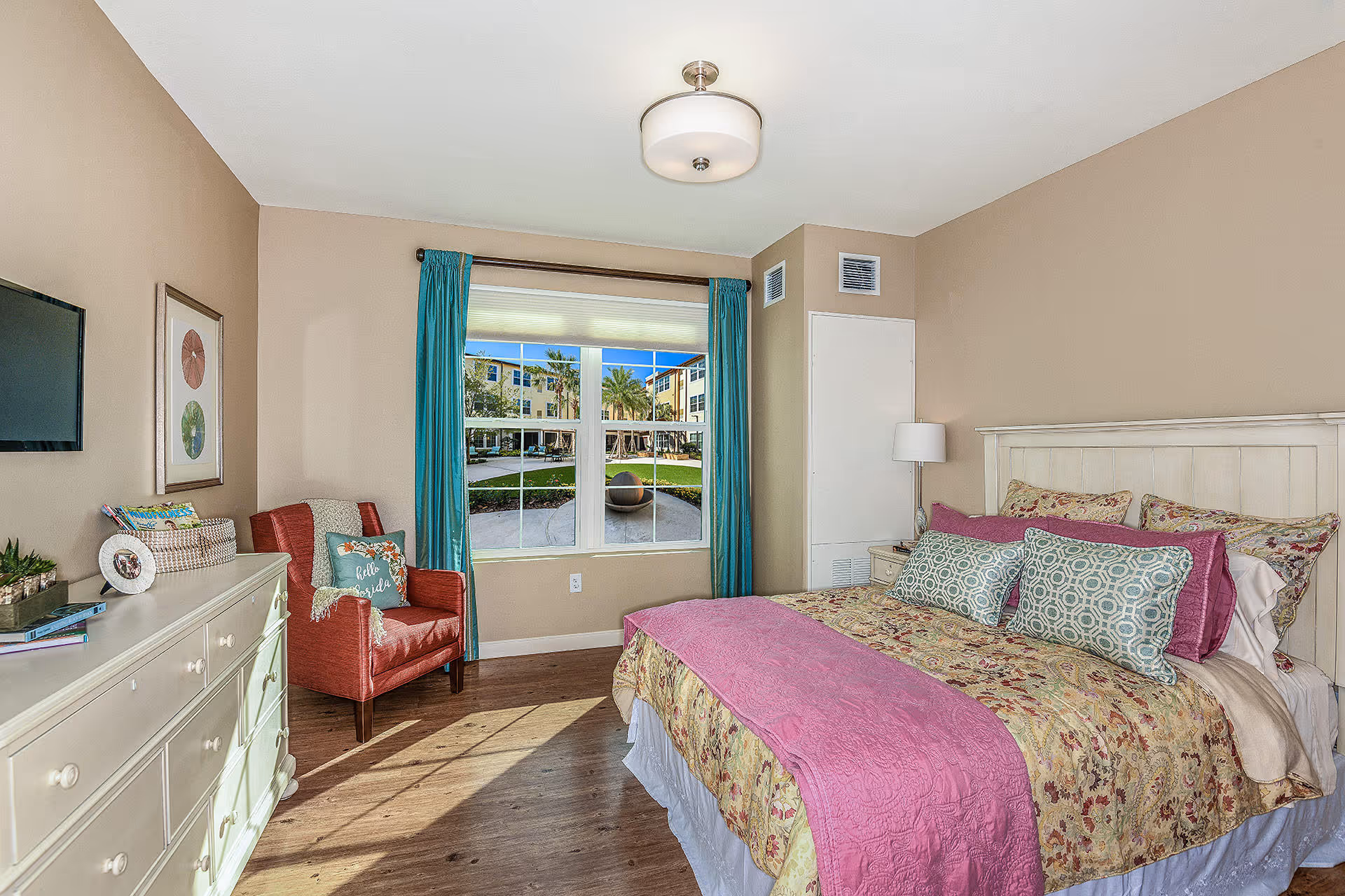 A cozy bedroom with a large bed covered in floral bedding and pink accents. There is a white dresser with decorative items and a wall-mounted TV on the left side. A red armchair with a pillow and throw blanket is positioned near a window with teal curtains, showing an outdoor courtyard with palm trees and buildings.