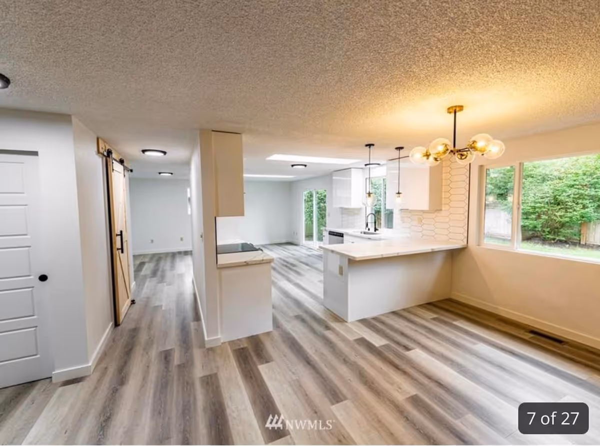Bright and modern open-concept interior space featuring a kitchen with white cabinetry, a white countertop island, pendant lights, and a unique chandelier. The room has large windows overlooking greenery outside, light wood flooring, and a sliding barn door on the left side.