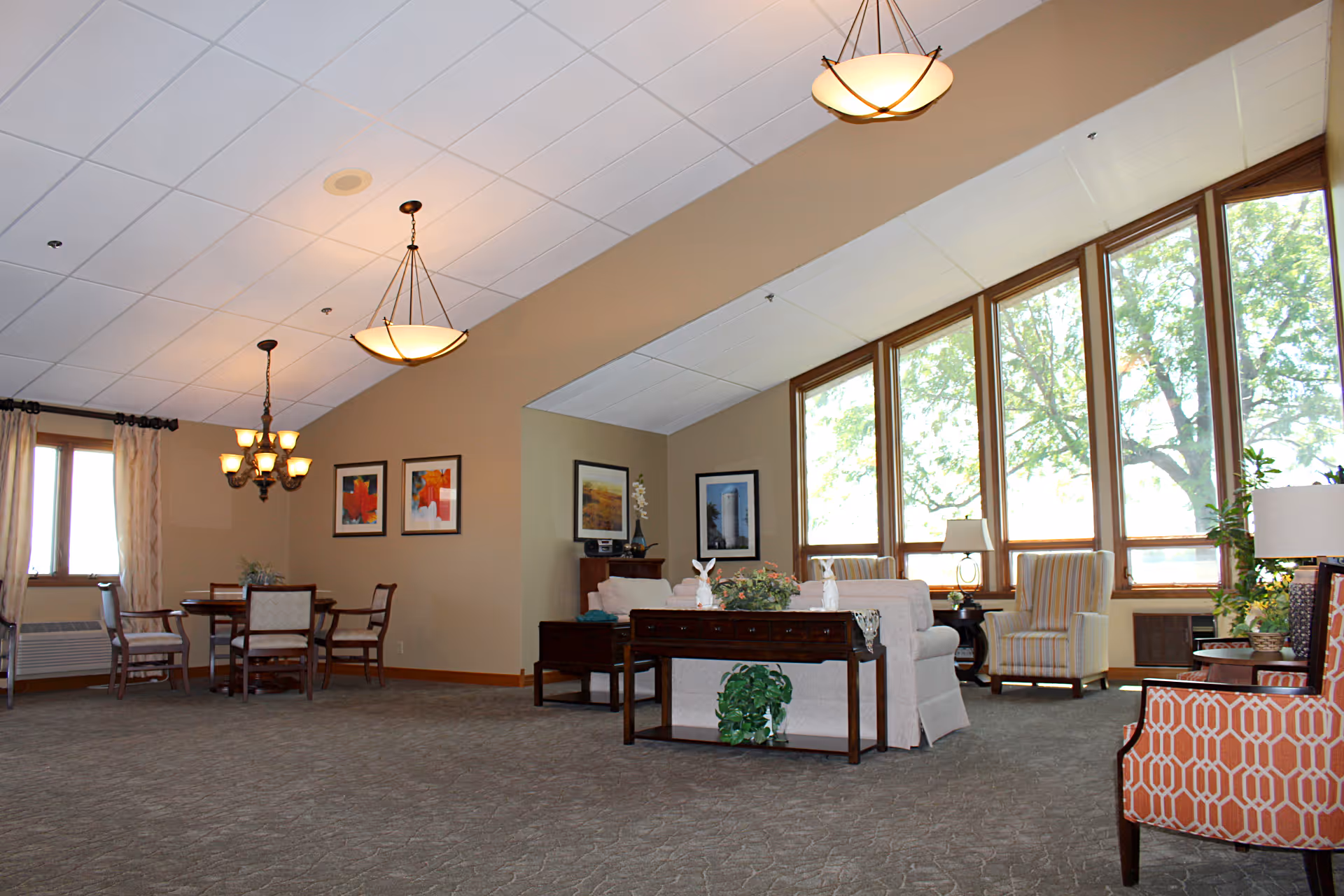Spacious senior living common room with sofas, chairs, a dining table, large windows and hanging light fixtures.