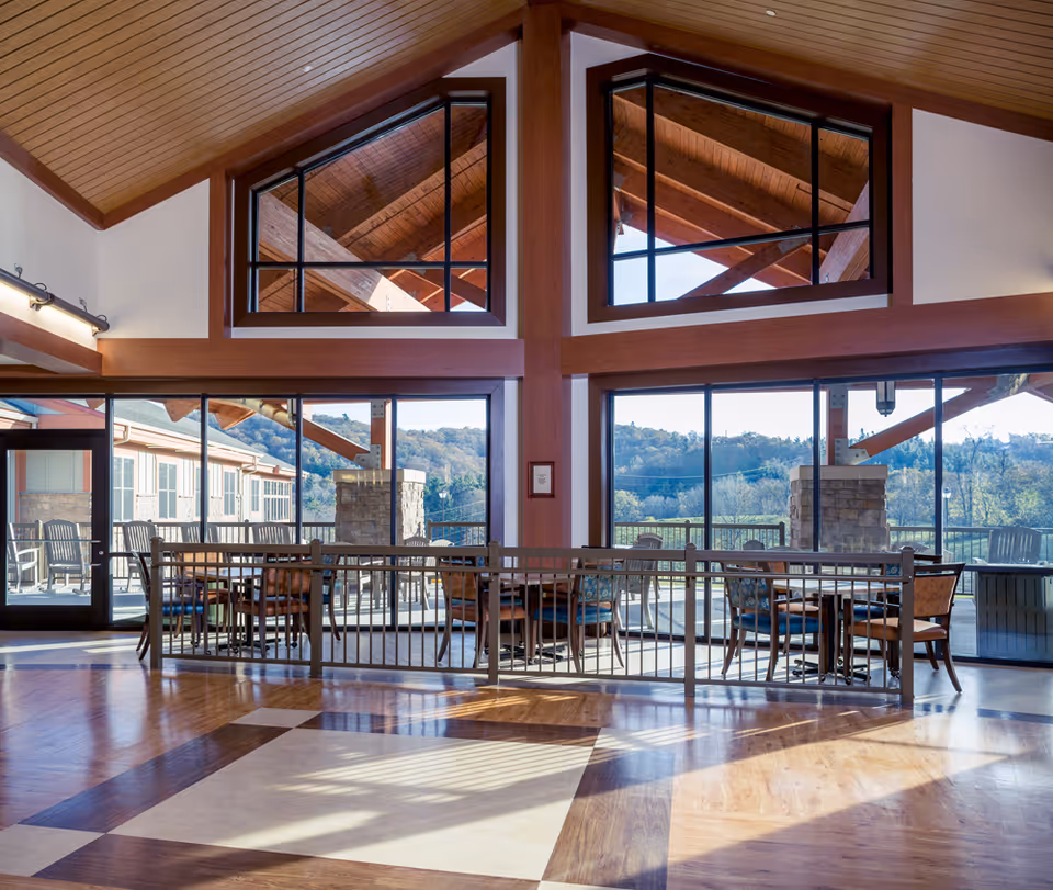 Spacious interior room with high wooden ceilings and large windows offering a view of an outdoor patio area with tables and chairs. The room features a polished wooden floor with a geometric pattern and a railing separating the seating area from the open space.