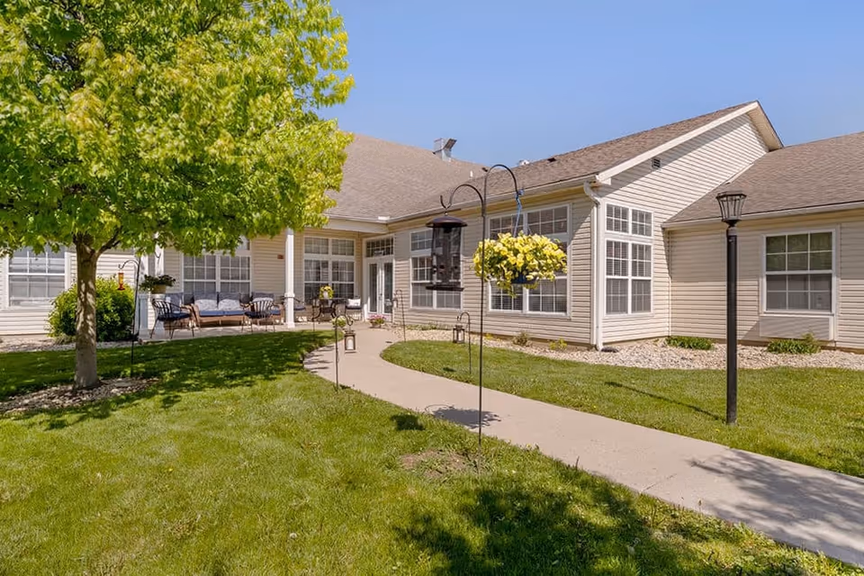 Outdoor view of a senior living facility with a paved walkway, green grass, a tree, hanging flower pots, and patio seating near the building under a clear blue sky.