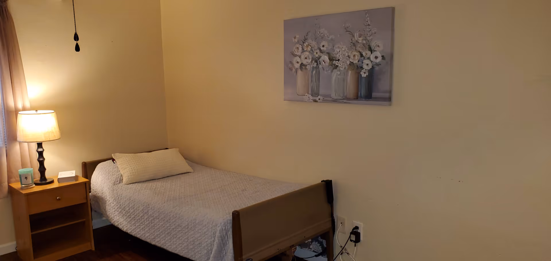 A simple bedroom with a single bed covered with a light gray quilt and a matching pillow. Next to the bed is a wooden nightstand with a lamp, a candle, and a small white device. A painting of white flowers in vases hangs on the beige wall above the bed. The room has wooden flooring and a window with beige curtains partially visible on the left.