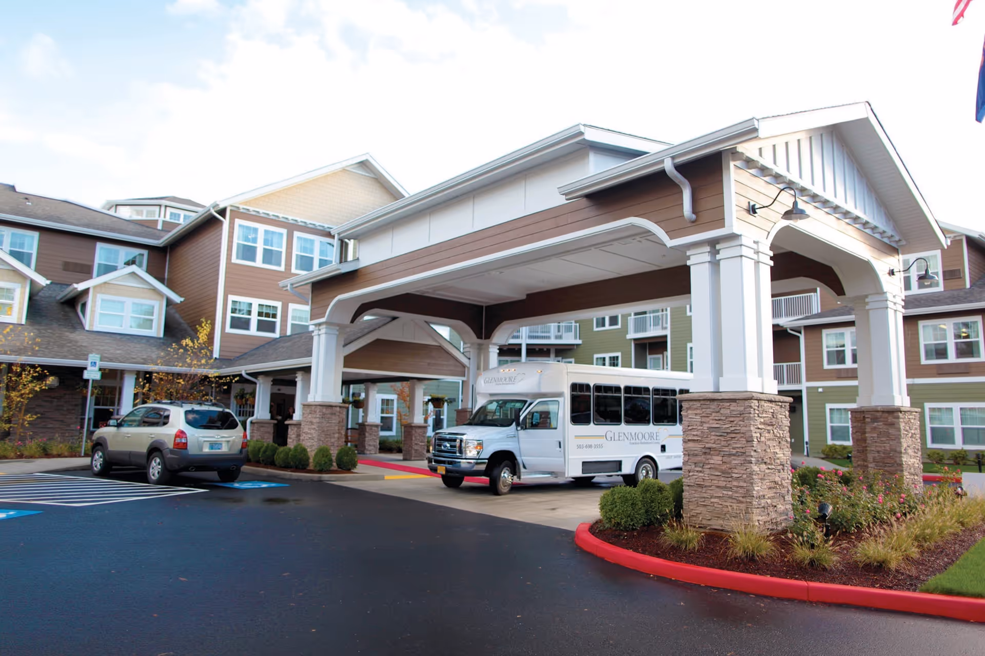 Exterior view of a senior living facility with a covered entrance supported by stone pillars. A white shuttle bus with the name Glenmoore is parked under the entrance, and a silver SUV is parked nearby. The building has multiple windows and a mix of brown and green siding with white trim. There are landscaped bushes and a red curb around the driveway.