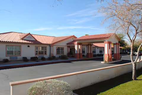 Exterior view of a single-story building with a red tiled roof and beige walls, featuring a covered entrance supported by columns. There is a driveway in front and some landscaping with bushes and a tree. The sky is clear and blue.