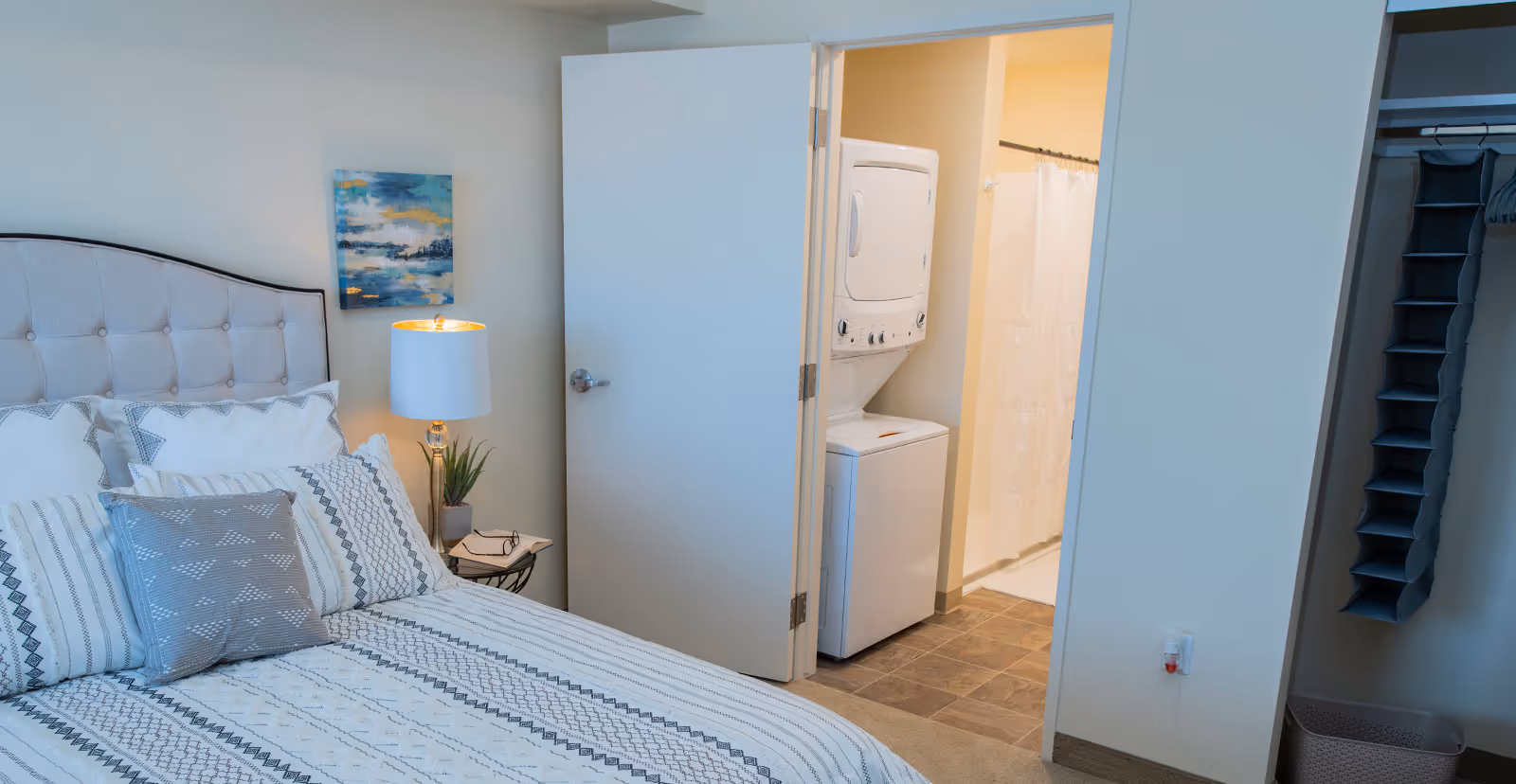 A bedroom with a neatly made bed featuring patterned bedding and multiple pillows. A bedside table with a lamp and a small plant is next to the bed. The door is open, revealing a laundry area with a stacked washer and dryer, and a bathroom with a shower curtain. There is also an open closet with hanging organizers and a laundry basket.