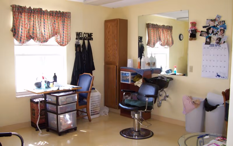 A small room with a window covered by a plaid valance, a wooden chair, a small table with various items, a black salon chair in front of a sink, a tall wooden cabinet, a large wall mirror, a calendar on the wall, and two trash bins. The room appears to be a personal grooming or hair washing area.
