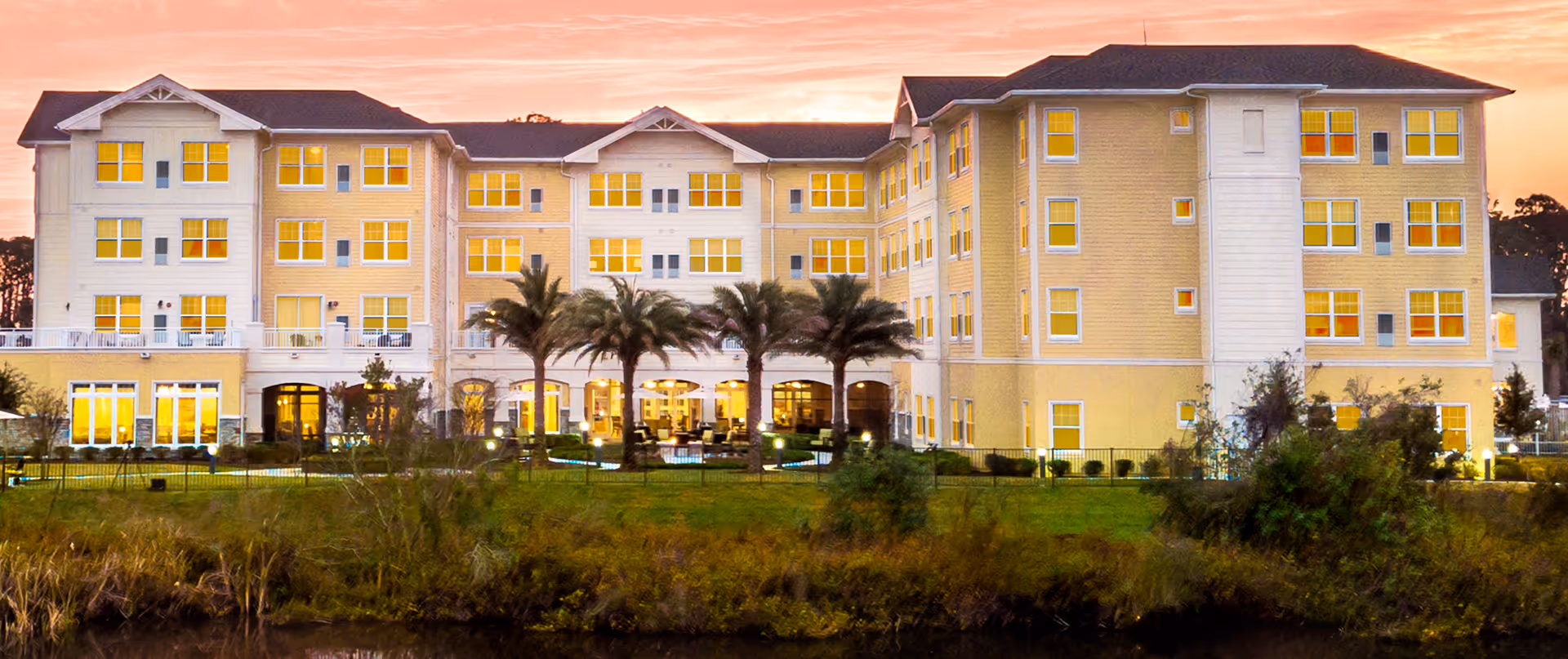 Exterior view of a large, multi-story senior living facility building named Sonata Windermere at sunset, with illuminated windows, palm trees, and a grassy area in the foreground.