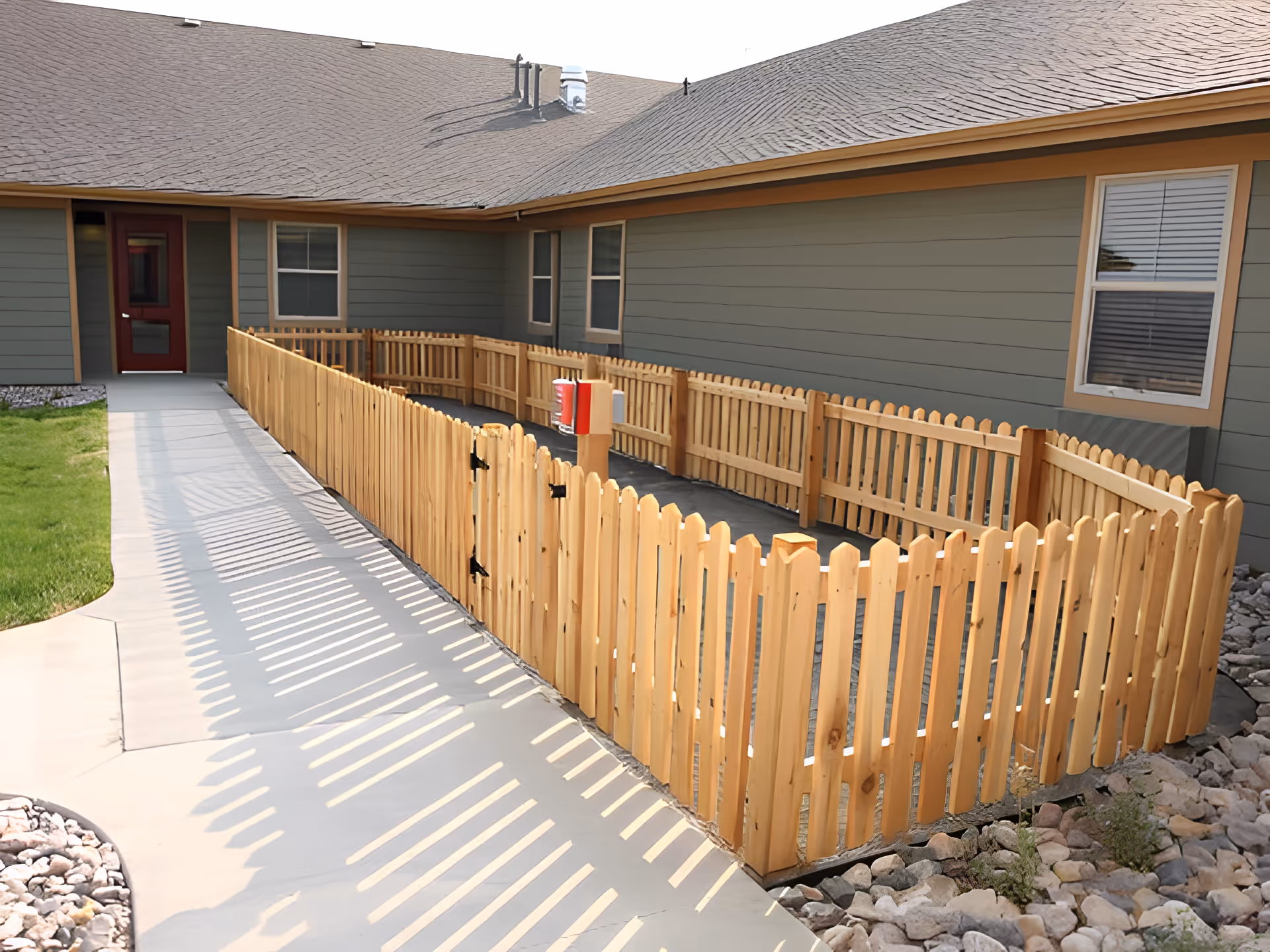 Outdoor area of Deer Trail Assisted Living featuring a wooden picket fence enclosing a small garden or patio space next to a building with gray siding and a brown roof. A concrete walkway runs alongside the fenced area, and there are windows and a red door on the building.