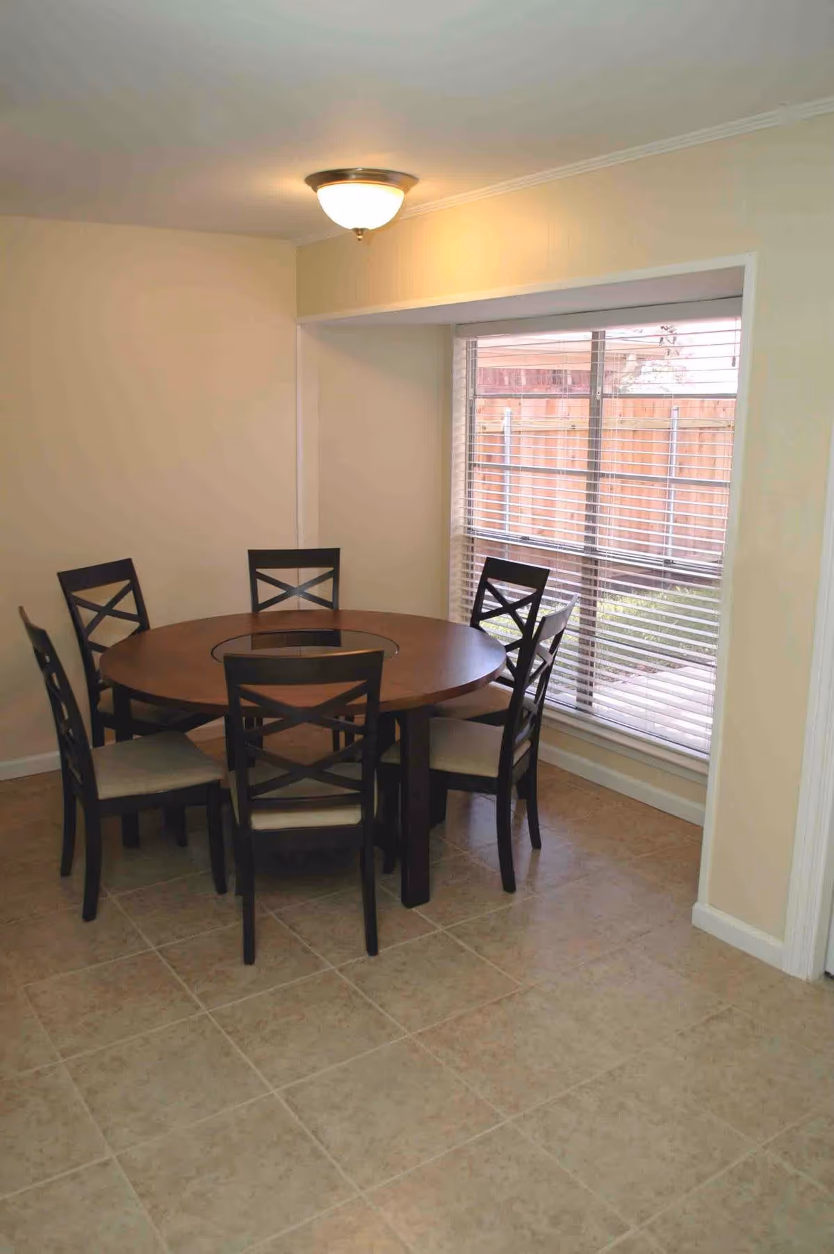A dining area with a round wooden table surrounded by six dark wooden chairs with beige cushions. The room has beige walls, a tiled floor, a ceiling light fixture, and a large window with white blinds overlooking a fenced backyard.