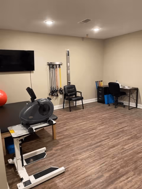 Small exercise/therapy room with a recumbent bike, resistance bands on the wall, a TV, chairs, and a desk.
