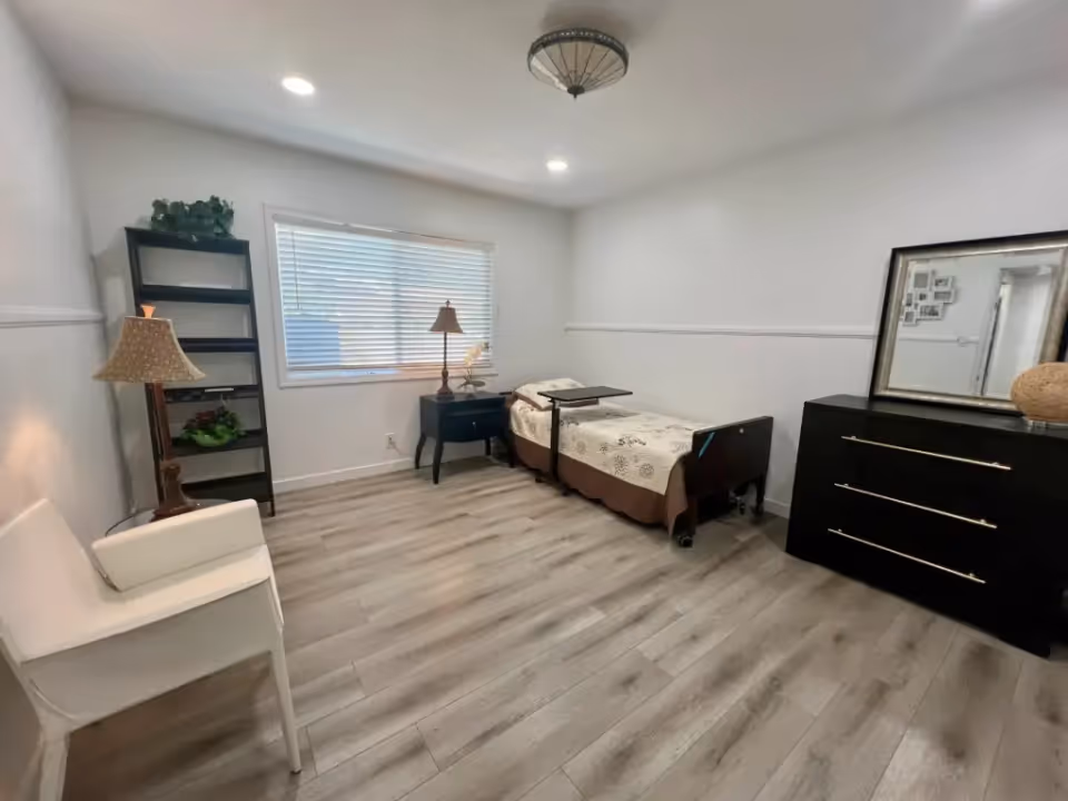 A simple, clean bedroom with a single bed covered with a patterned bedspread, a small black nightstand with a lamp, a black dresser with a large mirror, a white chair, a standing lamp, and a black shelving unit with plants. The room has light-colored wooden flooring and white walls with a window letting in natural light.