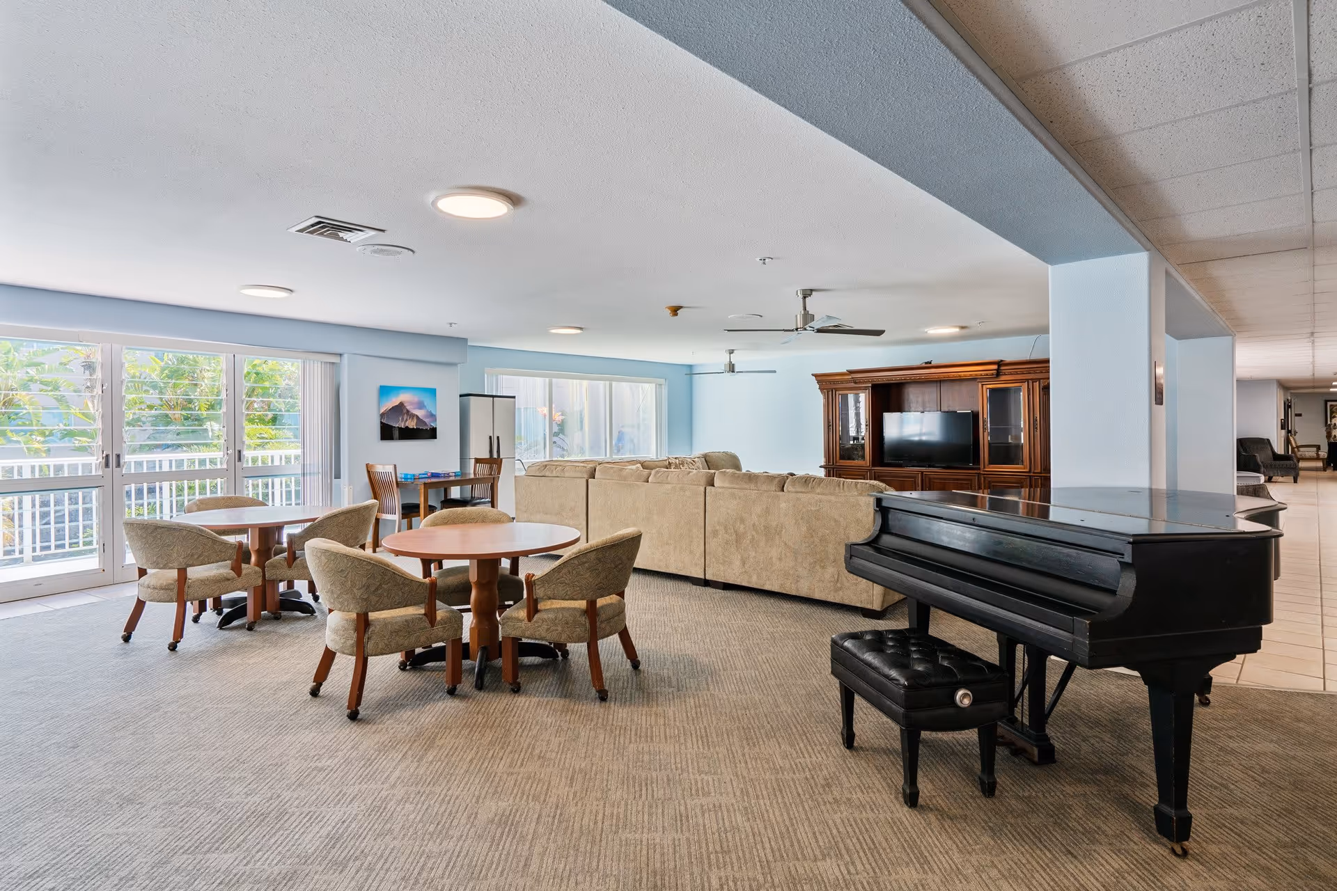 A spacious common area in a senior living facility featuring a black grand piano with a matching bench, several round tables with upholstered chairs, a large beige sectional sofa, a wooden entertainment center with a flat-screen TV, and large windows allowing natural light to fill the room.