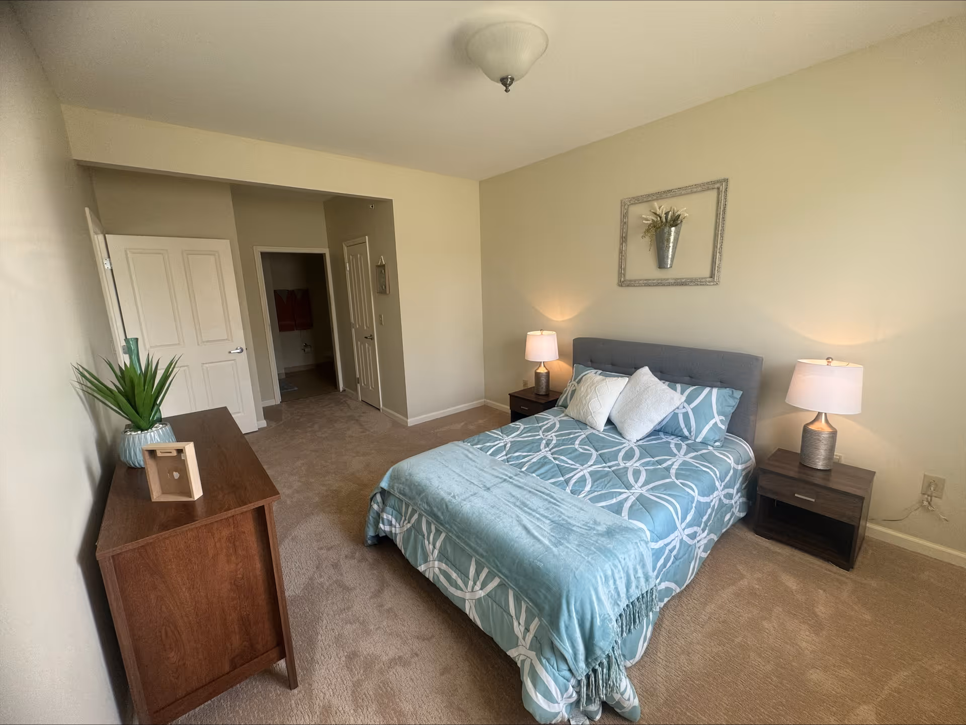 Bright, neutrally decorated bedroom featuring a bed with patterned teal bedding, two bedside tables with lamps, a wooden dresser, and an open doorway.
