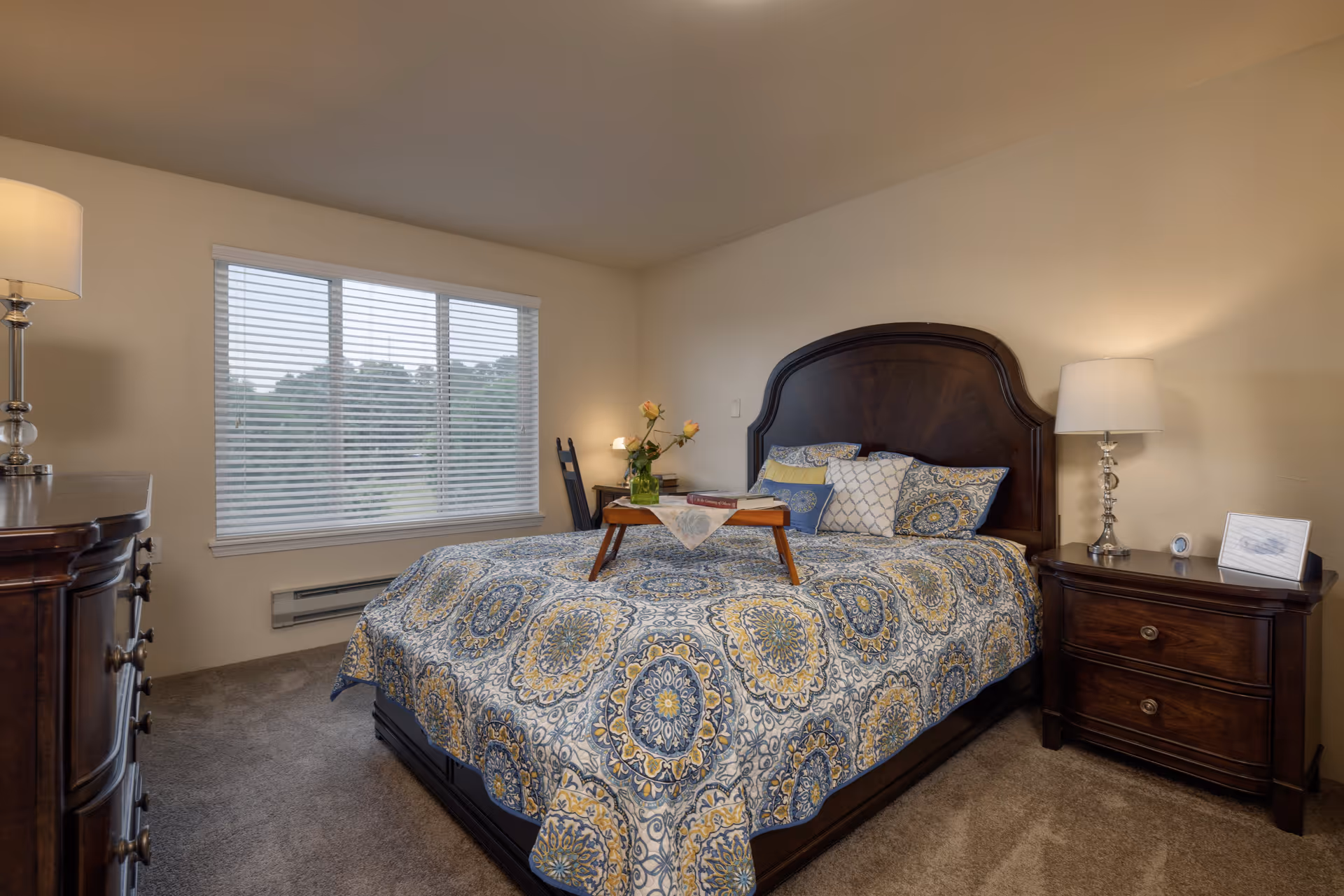 A cozy bedroom with a large bed featuring a patterned blue and yellow bedspread and multiple pillows. There is a wooden headboard and matching wooden nightstands on either side of the bed, each with a lamp. A window with white blinds lets in natural light, and a wooden dresser is visible on the left side of the room. A small tray with a book and flowers sits on the bed.