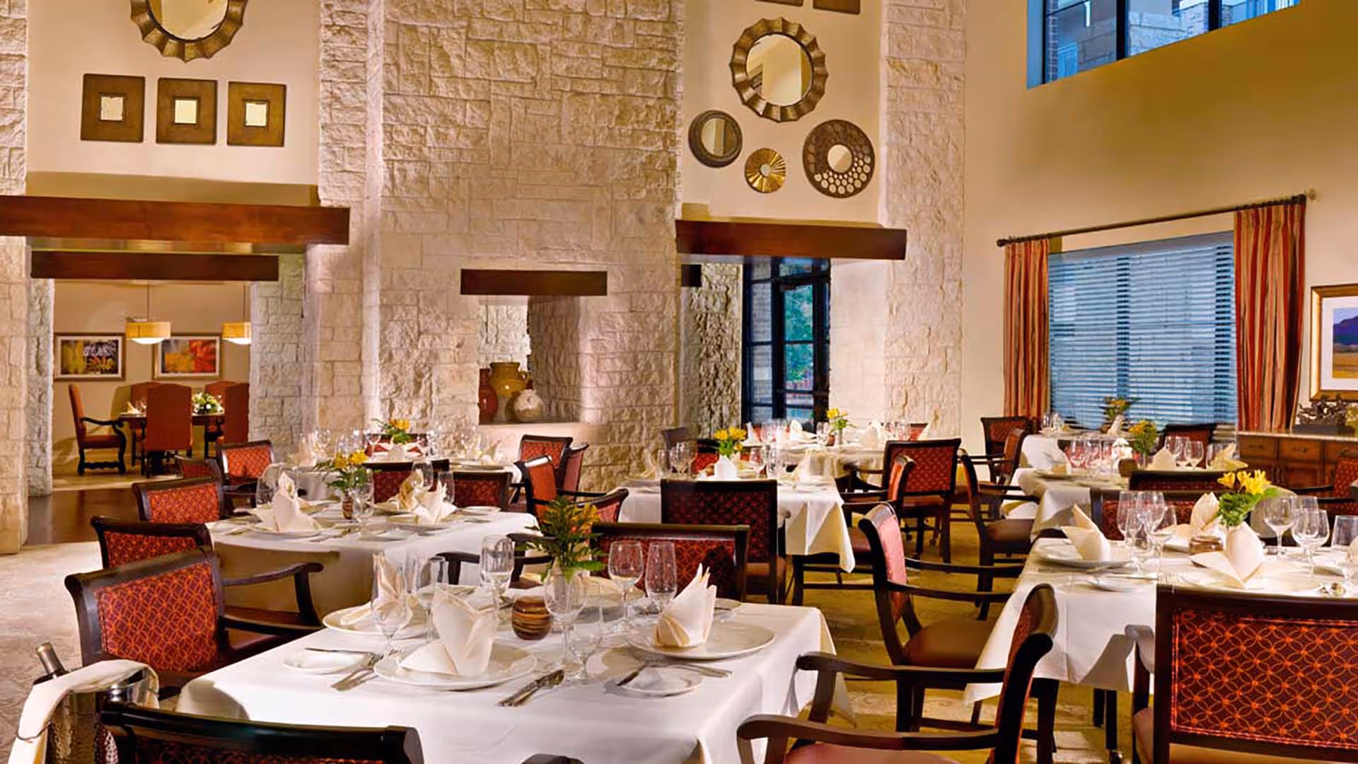 Elegant dining room with multiple tables set with white tablecloths, folded napkins, glassware, and silverware. The room features stone walls, decorative mirrors, large windows with striped curtains, and warm lighting. Chairs have red patterned upholstery.