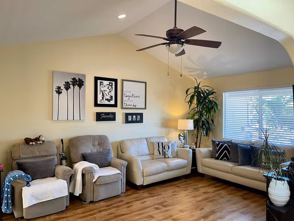 Bright living room with two beige sofas, two recliners, wall art, potted plants, a ceiling fan, and wood flooring.