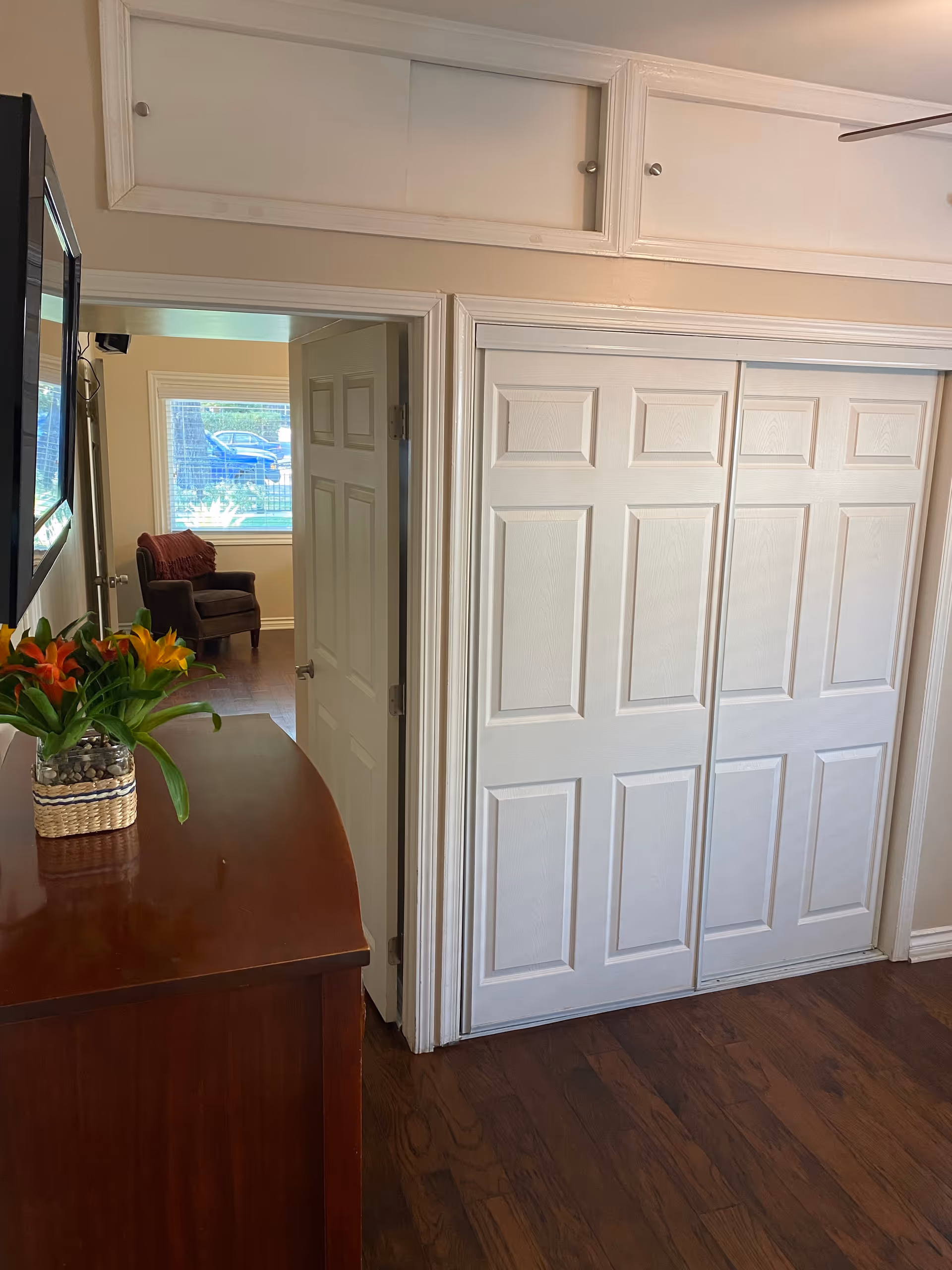 Interior view of a hallway showing white sliding closet doors, a wooden dresser with a vase of flowers, and a doorway leading to a room with a chair.