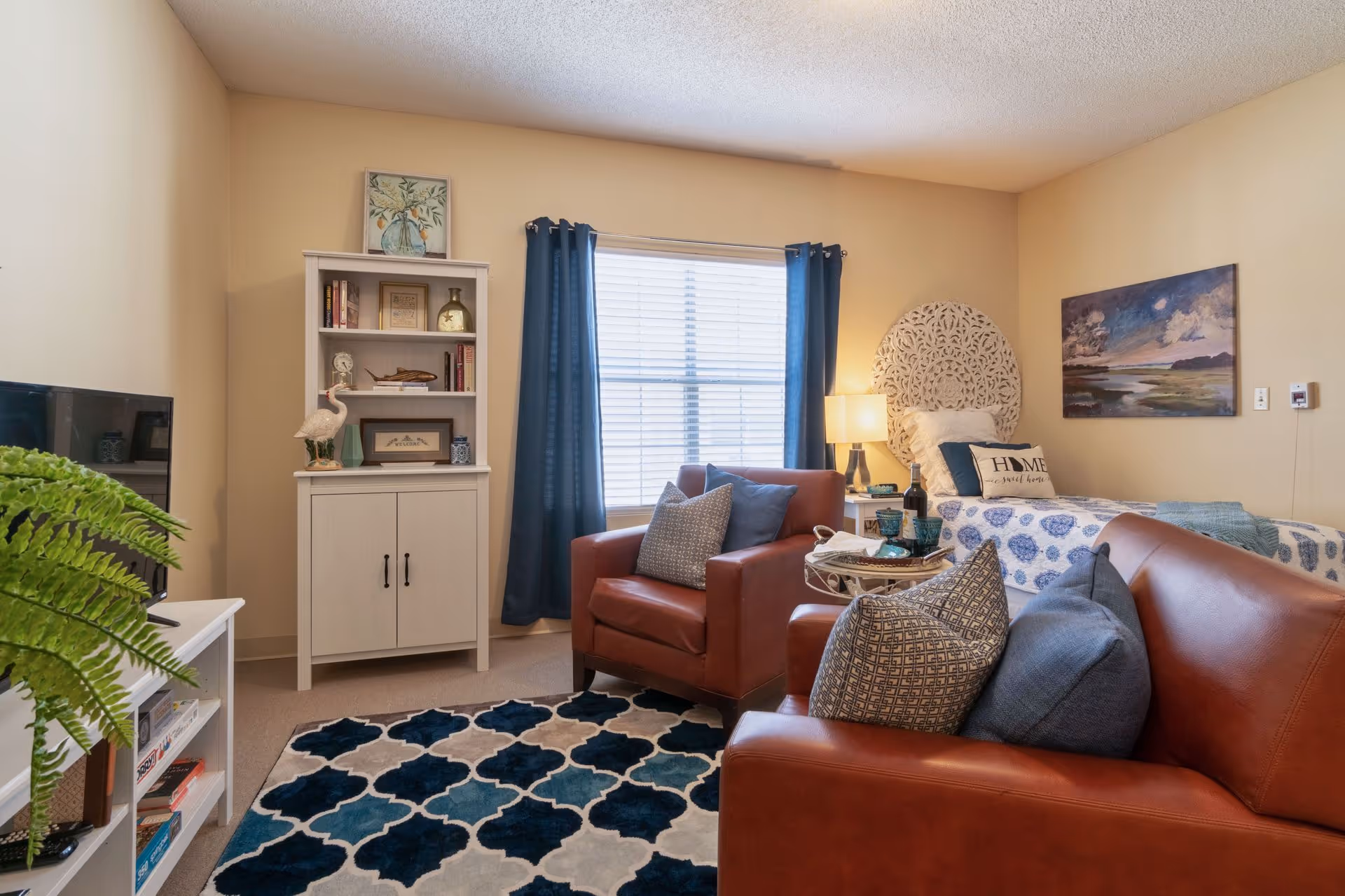 A cozy senior living room with a single bed featuring a decorative headboard and blue patterned bedding. The room includes a brown leather armchair and sofa with patterned cushions, a white cabinet with shelves holding books and decorative items, a window with blue curtains, a table lamp, a framed painting on the wall, a patterned area rug, and a television on a white stand with a green plant beside it.