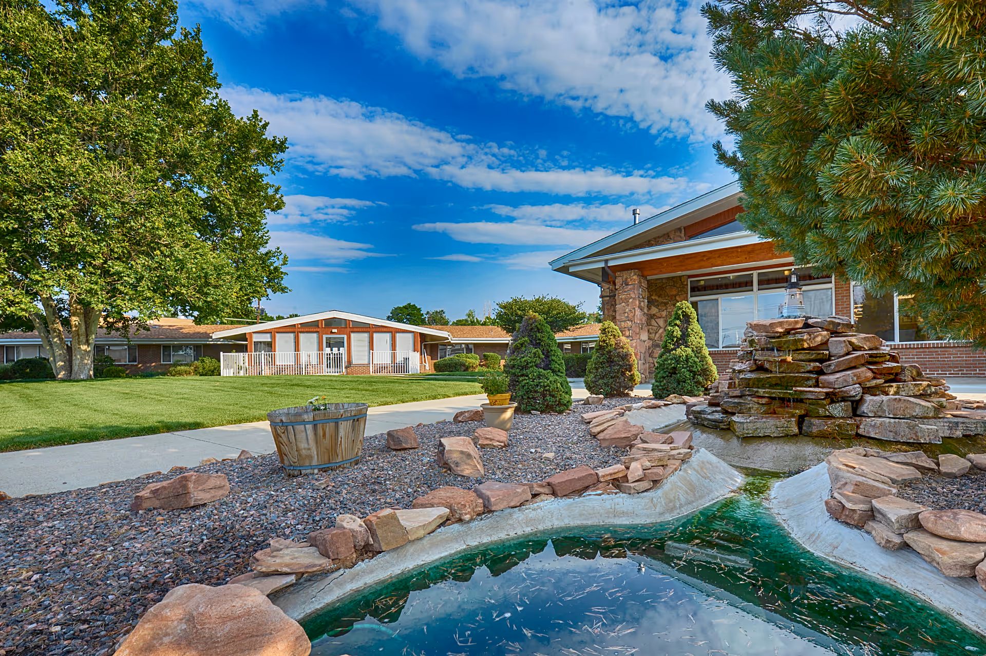 Outdoor view of Harmony Pointe Nursing Center featuring a landscaped garden with a small pond and rock waterfall, surrounded by trees, shrubs, and a well-maintained lawn under a partly cloudy blue sky.
