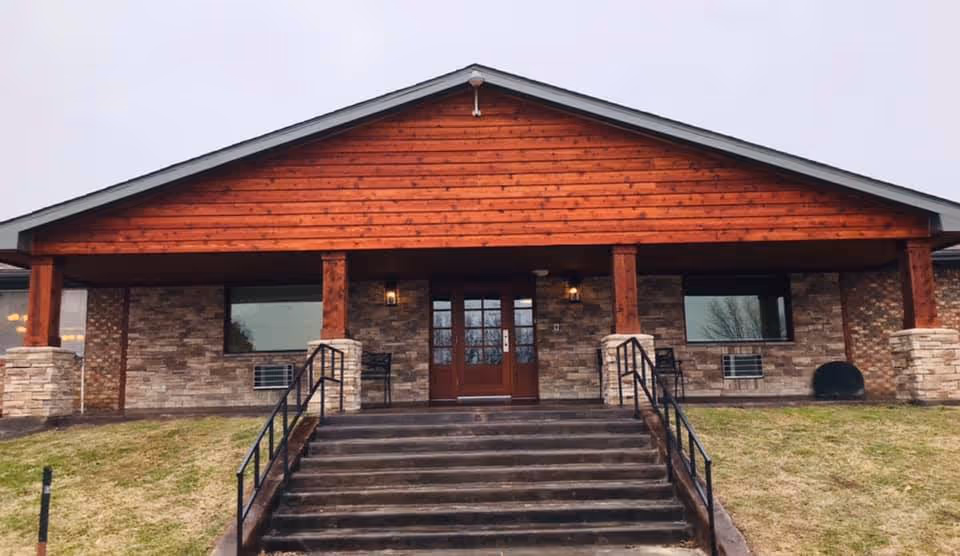 Front exterior view of a single-story building with a wooden gabled roof and stone facade. There are steps leading up to double wooden doors with glass panels, flanked by two windows and outdoor chairs on either side. The building is surrounded by a grassy area.