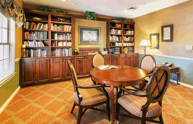 Cozy communal library/reading room with wooden bookshelves, a round table and four upholstered chairs.