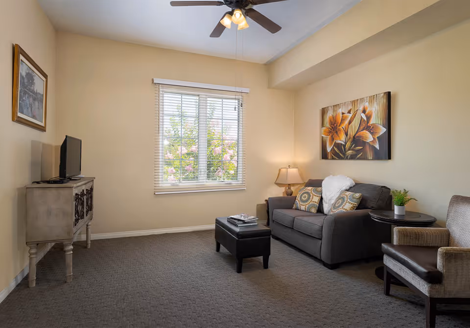 Furnished living room with a sofa, armchair, ottoman coffee table, TV on a console, ceiling fan, and a window with blinds.