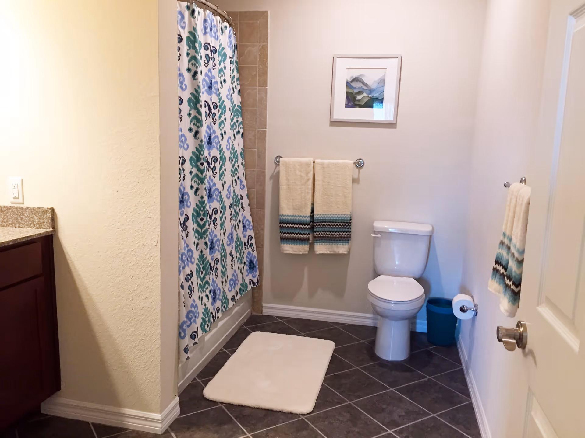A clean bathroom featuring a white toilet, a shower with a colorful blue and green patterned curtain, two beige towels with blue and black stripes hanging on a towel rack, a white bath mat on the tiled floor, a small blue trash bin, a framed abstract artwork on the wall, and a wooden vanity with a granite countertop partially visible on the left.