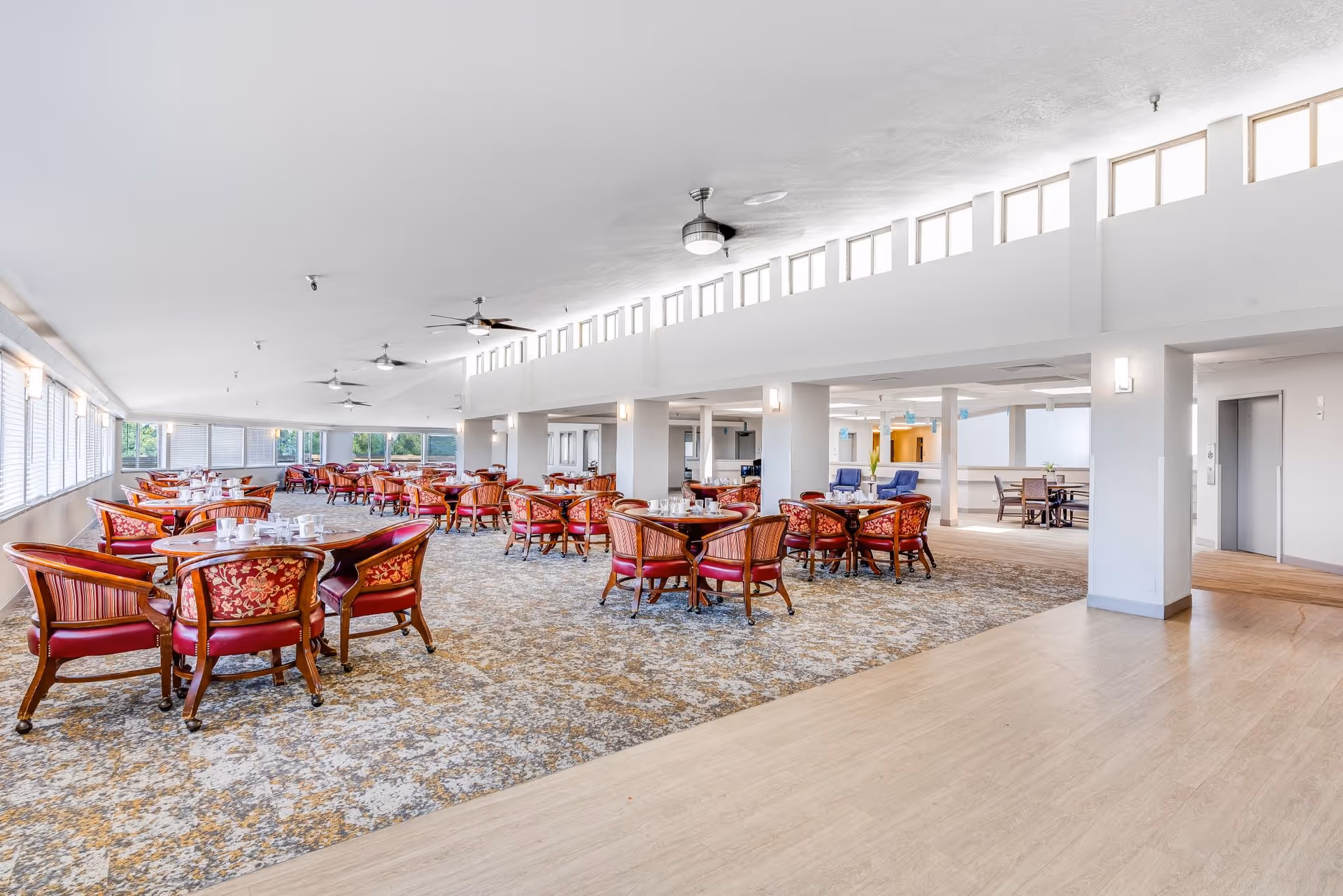 A spacious dining room with multiple round wooden tables surrounded by red cushioned chairs with floral patterns. The room has large windows on one side allowing natural light to fill the space. Ceiling fans and modern light fixtures are mounted on the white ceiling. The floor is a combination of patterned carpet and light wood flooring.