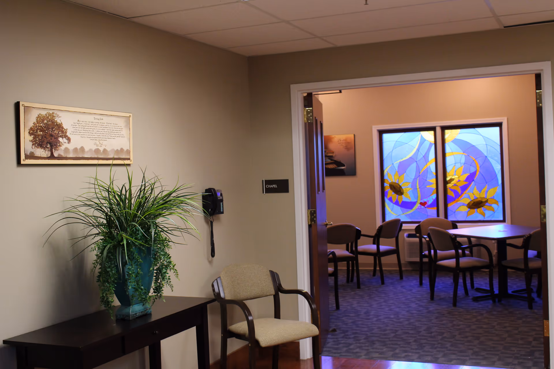 Interior view of a hallway leading to a chapel room in a memory care facility. The hallway has a beige wall with a framed inspirational quote and a green potted plant on a dark wooden table. A beige cushioned chair is placed next to the table. The chapel room has an open door, several chairs around tables, and a stained glass window with a sunflower design.