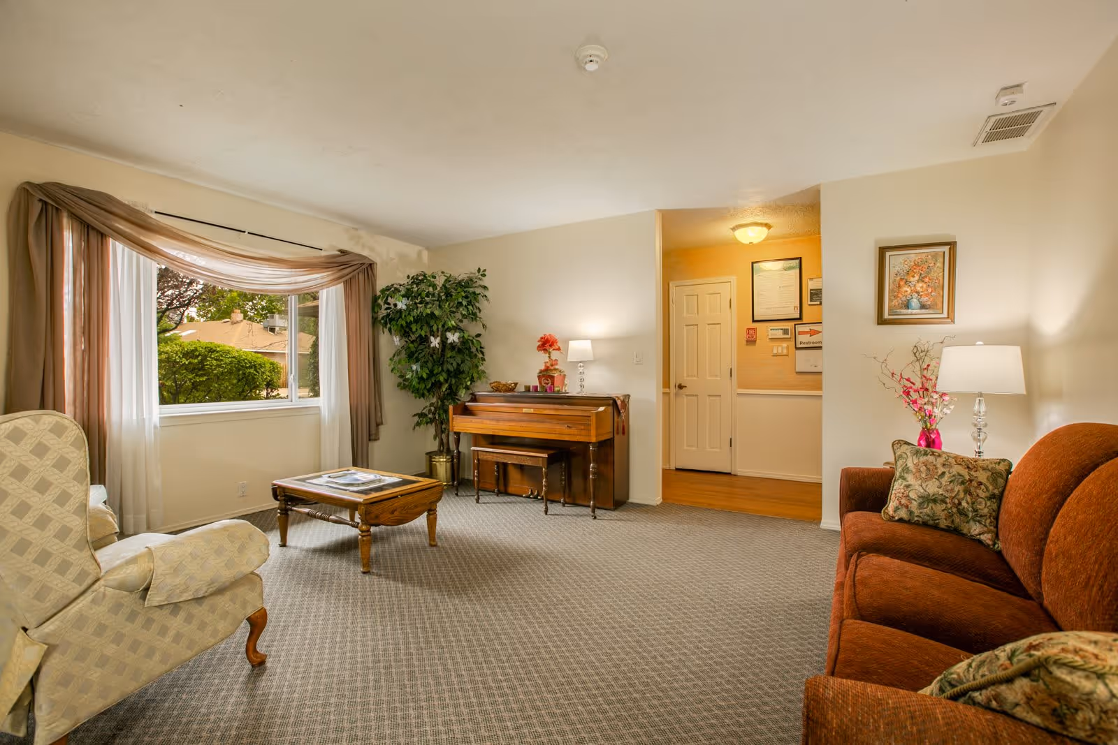 A cozy living room with a large window draped with sheer and brown curtains, a beige armchair, a wooden coffee table, a brown sofa with floral cushions, a piano against the wall, a potted plant, and two table lamps providing warm lighting. There is a framed picture on the wall and a doorway leading to another room.