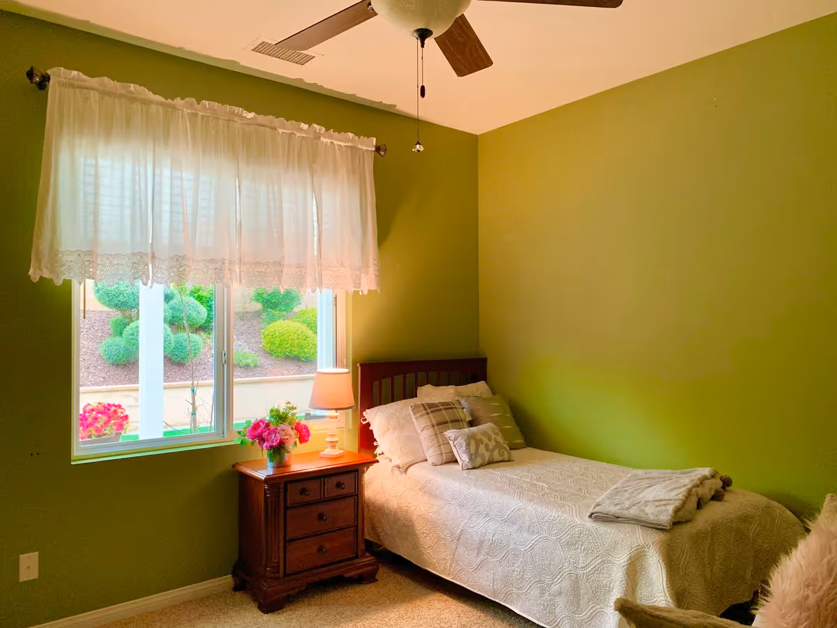 Small bedroom with a single bed against a green wall, a wooden nightstand with a lamp and flowers, and a window with lace curtains.