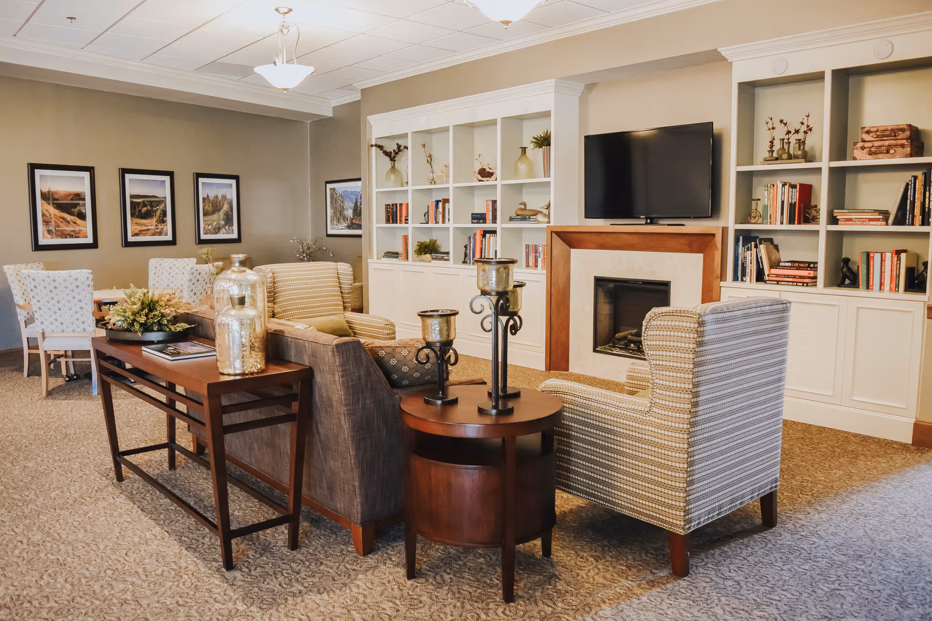 A cozy senior living common area with a seating arrangement including a sofa and armchairs around a small round wooden table with candle holders. Behind the seating area is a built-in white bookshelf filled with books and decorative items, and a flat-screen TV mounted above a fireplace. On the left side, there is a dining area with a table and chairs, and framed landscape pictures on the wall.