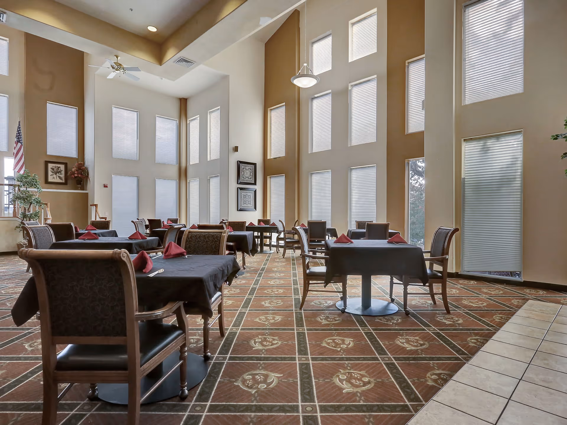 A spacious dining room with high ceilings and multiple tall windows covered with blinds. Several tables are set with black tablecloths and red folded napkins, surrounded by wooden chairs with cushioned seats. The room features patterned carpet flooring and a tiled area near the entrance. There are framed pictures on the walls and an American flag in the corner.