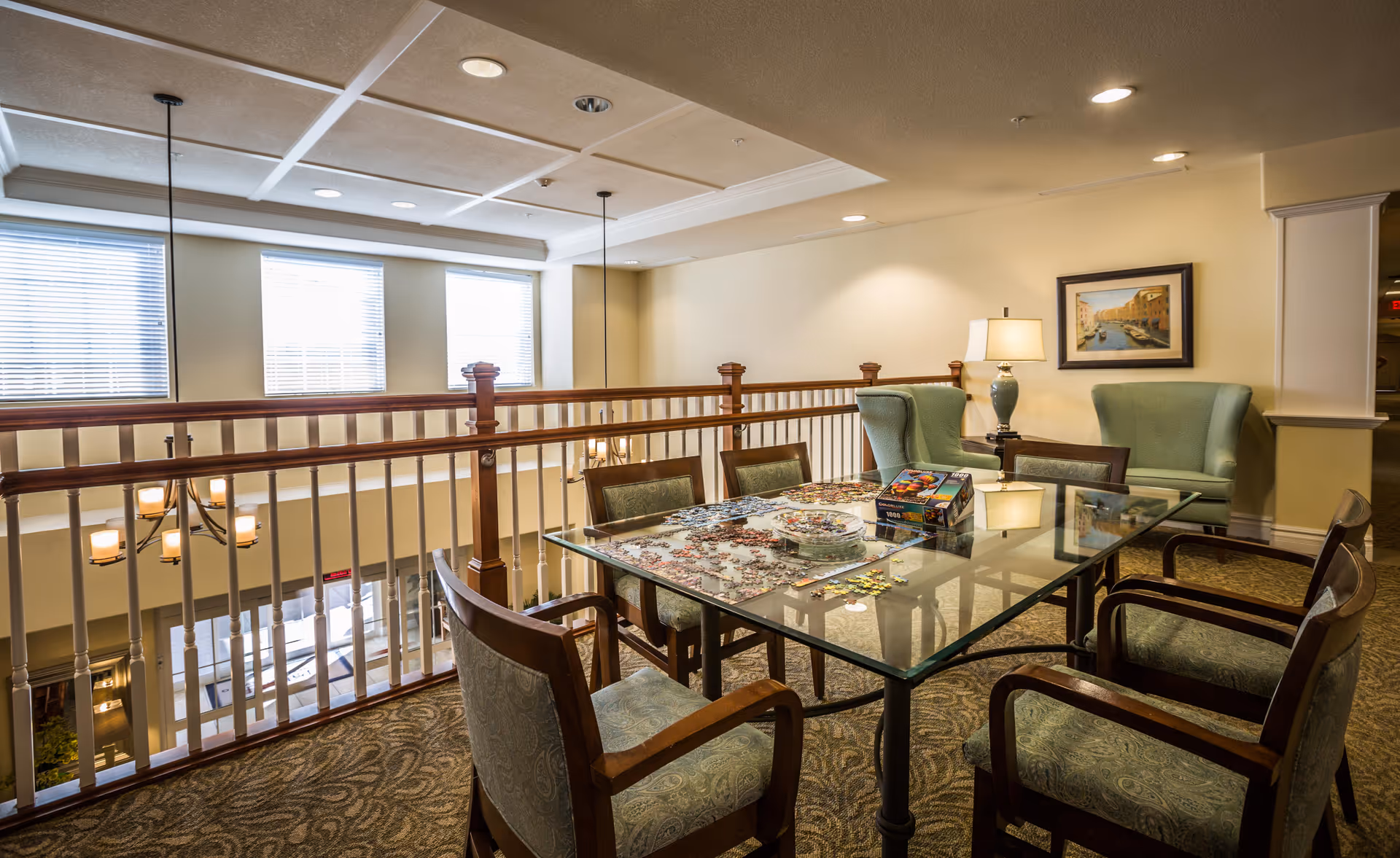 A cozy senior living common area with a glass table in the center, surrounded by six cushioned chairs. On the table, there is a partially completed jigsaw puzzle and a puzzle box. In the background, two green armchairs flank a side table with a lamp and a framed painting on the wall. The area overlooks a lower floor through a wooden railing, with large windows letting in natural light.